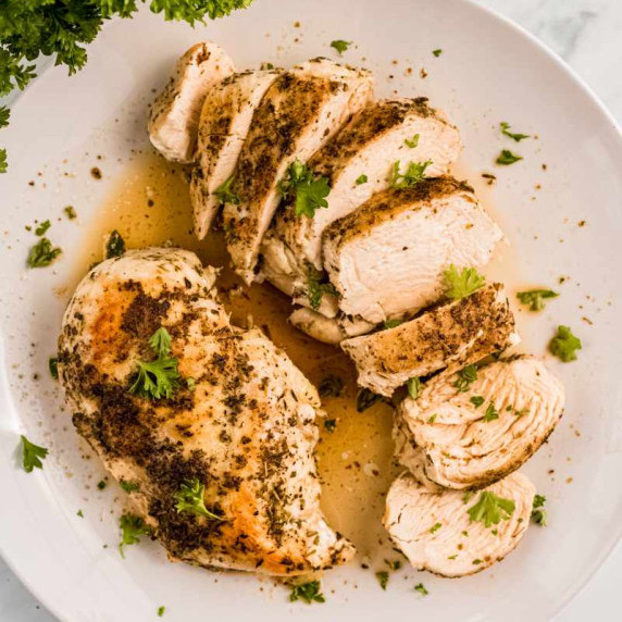 Two seasoned chicken breasts, one whole and one sliced, on a white plate with flavorful juices.