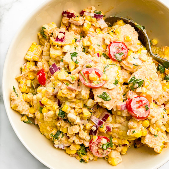 A white bowl filled with corn and tomato salad, asppon resting in the salad to the side in the bowl.
