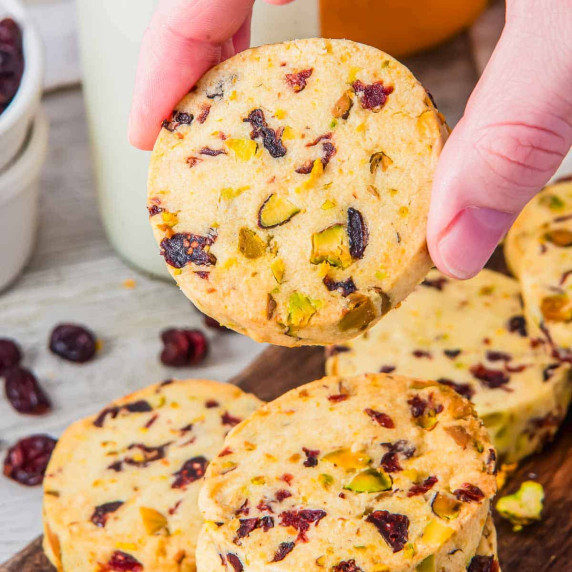 Hand holds a shortbread cookie with dried cranberries and pistachios. More cookies on a board.
