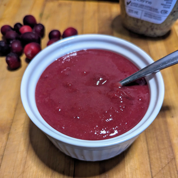 A bowl of cranberry mustard dipping sauce on a board with a spoon and fresh cranberries.