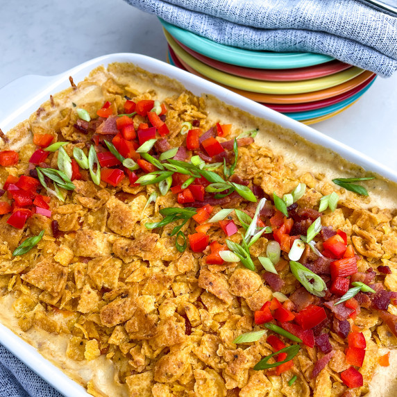 White casserole dish with corn casserole topped with Fritos, green onions, red pepper and bacon. 