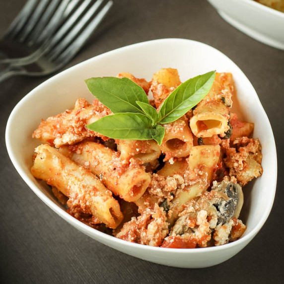 Close up shot of a white bowl with baked ziti garnished with fresh basil on a brown surface.