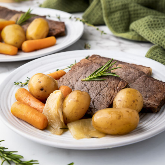Side shot of rump roast, potatoes, and carrots on a white plate garnished with rosemary.