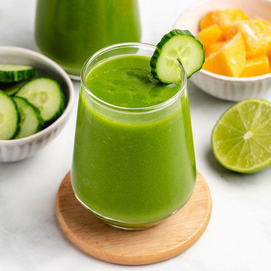 Mango cucumber smoothie in a glass on a coaster with a cucumber slice.