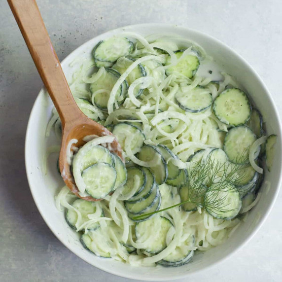 A wooden spoon scoops some cucumber onion salad out of a large white serving bowl.