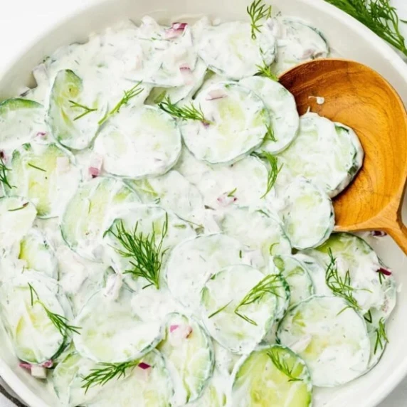 creamy cucumber onion salad topped with chopped fresh dill in a bowl with a wooden spoon