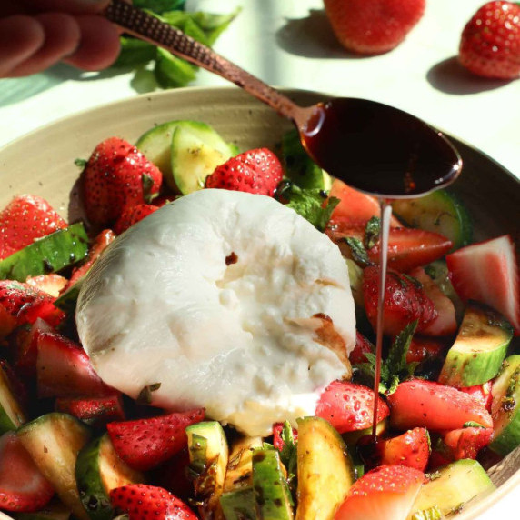 Balsamic glaze being drizzled over a cucumber strawberry salad with burrata.