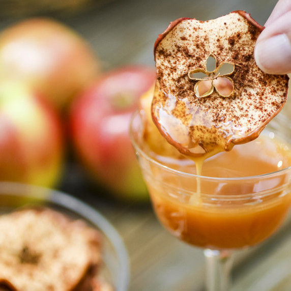 Side close up shot of a hand dipping a cinnamon apple chip into a glass with caramel.