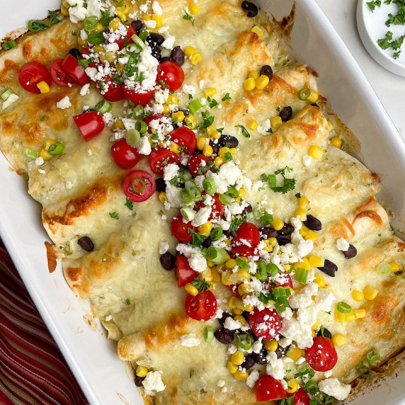 White casserole dish with chicken enchiladas and tomatoes, corn, black beans, feta, and green onions