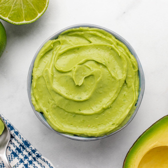 Vegan avocado lime crema in a bowl on table with a sliced lime and avocado.