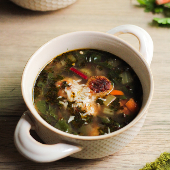 Easy Italian wedding soup in a beige bowl with handles, on a light wooden surface.