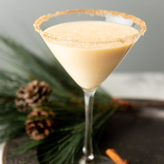 A creamy eggnog martini in a martini glass rimmed with cinnamon and sugar stands on a serving tray.