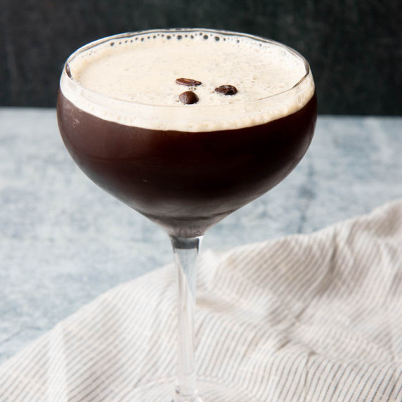 A coupe glass filled with an espresso martini garnished with coffee beans stands on a kitchen linen.