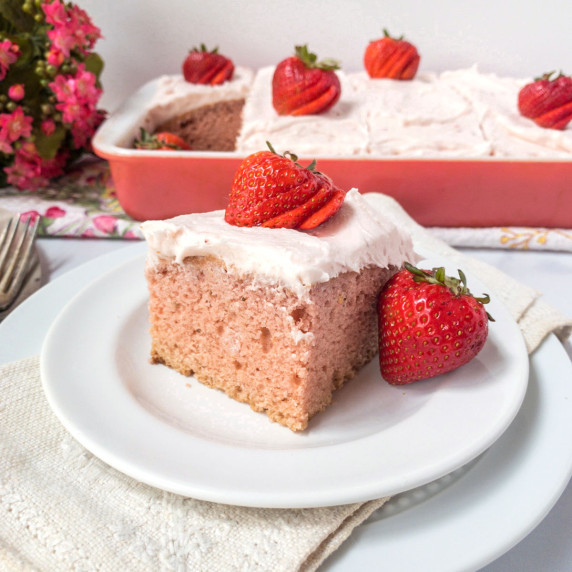 A slice of strawberry cake on a white place with a sheet cake in the background.