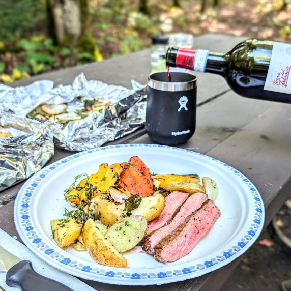 Campfire grilled flank steak on a paper plate on a camping table with wine being poured behind it