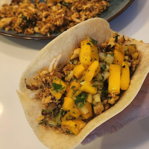 A hand holds a savory taco filled with diced mango, fresh herbs, and a mix of cooked fish