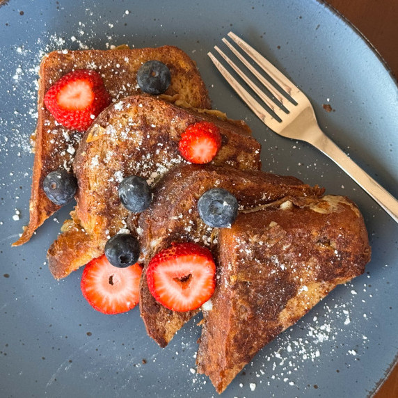 Four pieces of French Toast on a blue plate with blueberries and strawberries and a fork beside it