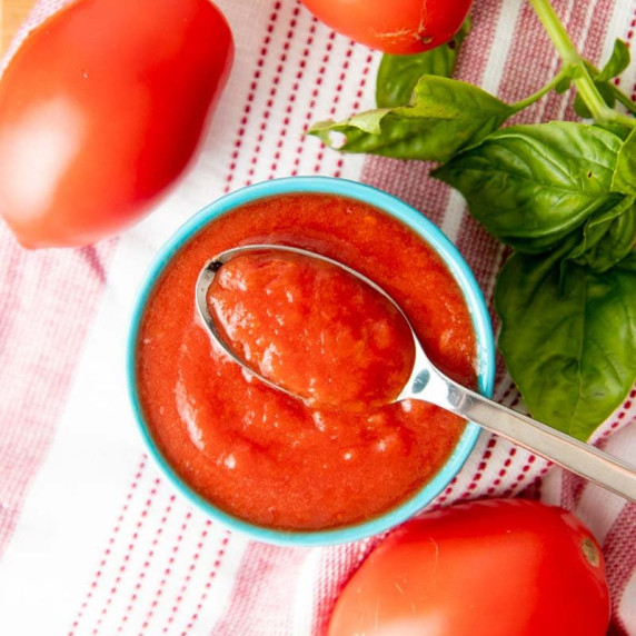 A spoonful of tomato puree sits atop a bowl of homemade puree, fresh basil and tomatoes around it.