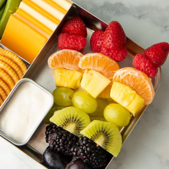 Four rainbow fruit skewers in a bento style lunch box with yogurt dip, cheese and crackers, and veg.