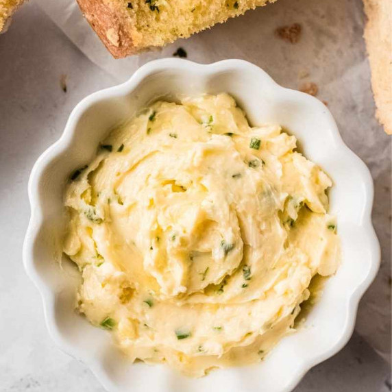 A scalloped white ramekin filled with homemade garlic herb butter with pieces of garlic bread nearby