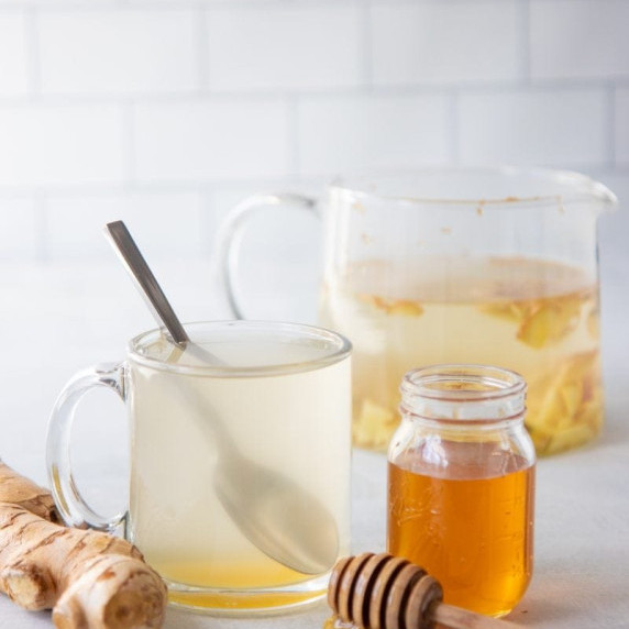 A clear mug of ginger tea with a spoonful of honey sits in front of a large glass pitcher of tea.
