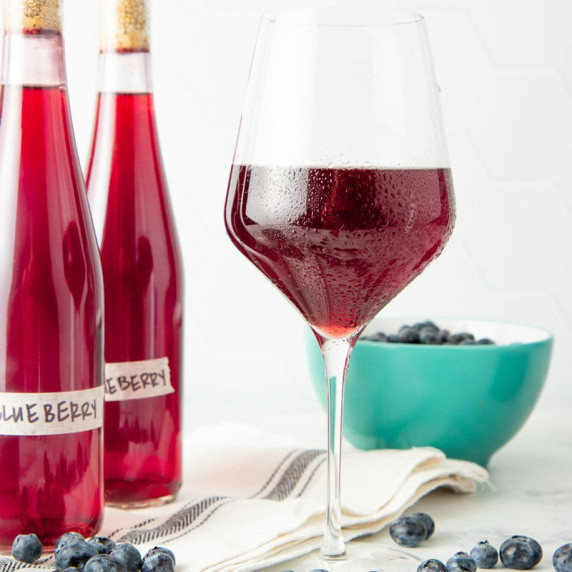 A chilled glass of blueberry wine stands before two bottles of wine and a bowl of fresh blueberries.