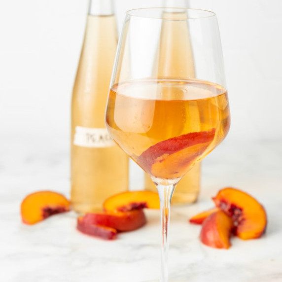 A glass of peach wine stands before two bottles of homemade wine and fresh peach slices.