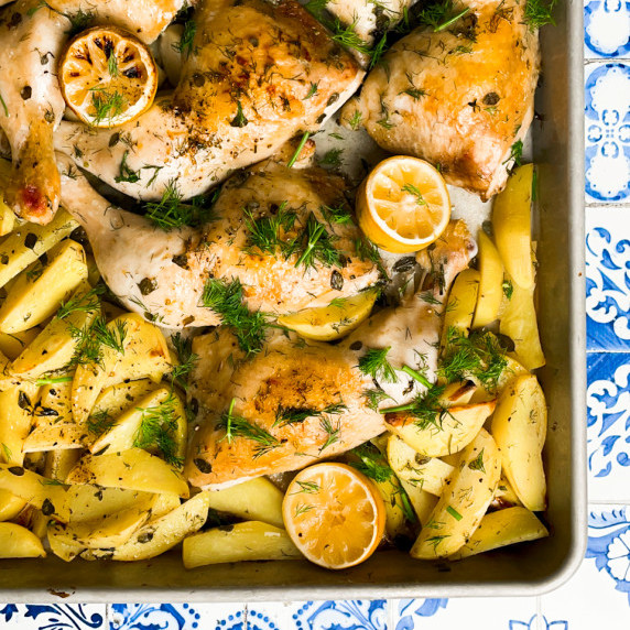 Chicken legs on a bed of potato wedges in a large metal sheet pan. Herbs covering the food.