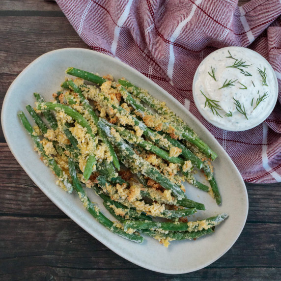 Green beans coated in panko on a serving plate next to a small bowl of homemade dip