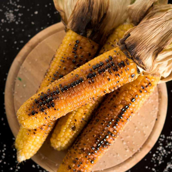 Grilled corn on the cob piled onto a wooden serving platter with charred husks pulled back and salt.