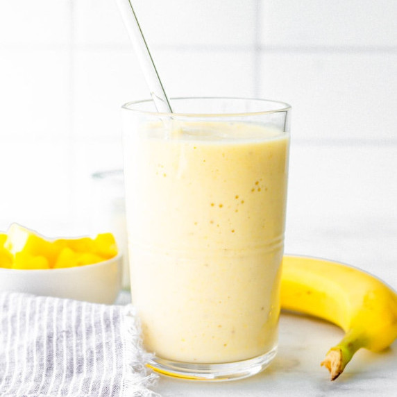 Coconut pineapple smoothie in a glass with a bowl of pineapple chunks and a fresh banana around it.