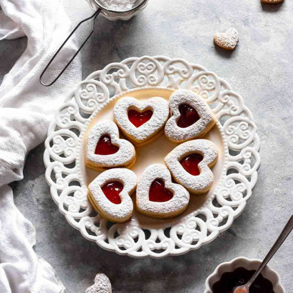 Heart jam cookies on a white plate next to powdered sugar and jam.