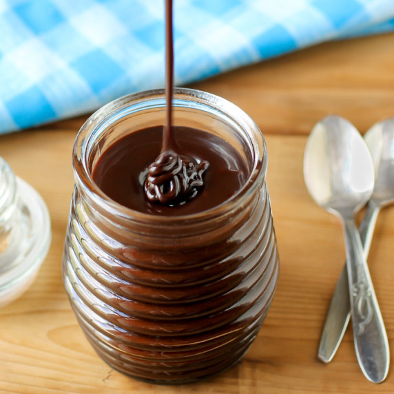 Close up shot of homemade chocolate sauce being drizzled into a clear jar with spoons on the side. 