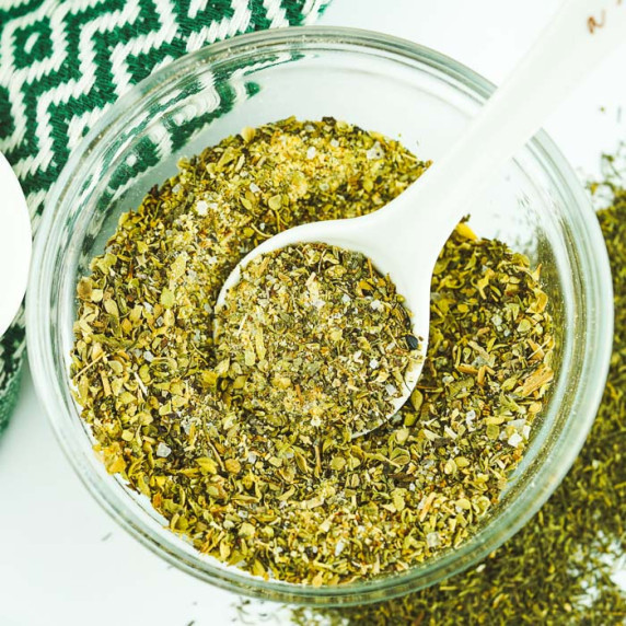 Overhead shot of greek spice mix in a clear jar with a white spoon inside on a white surface.