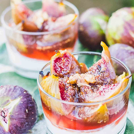 Honey roasted figs over Greek yogurt in small glasses next to fresh figs and leaves.