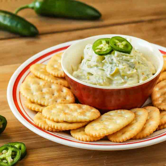 Side view of jalapeno artichoke dip in a red bowl surrounded by a ring of crackers on a red and whit
