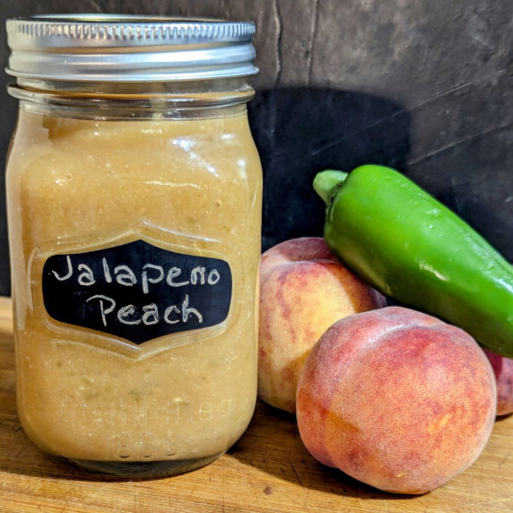 a jar of jalapeno peach bbq sauce with peaches and jalapeno on the side.