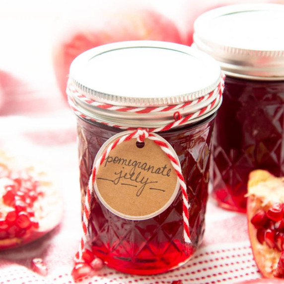 A jar of homemade pomegranate jelly with handmade gift tag stands amid open pomegranate sections.