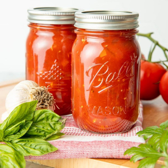 Two mason jars of canned homemade spaghetti sauce stand on a counter with fresh ingredients nearby.