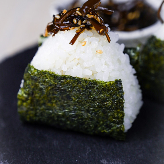 An onigiri rice ball wrapped with nori and topped with simmered kelp served on a slate plate