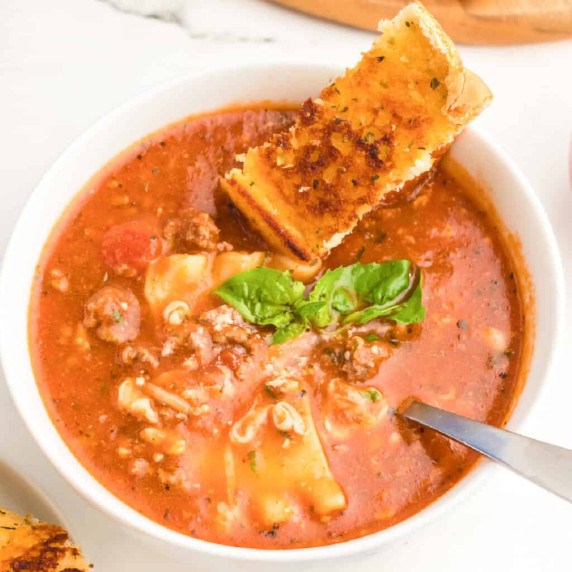 Lasagna soup in a bowl with a spoon topped with mozzarella cheese, basil and a slice of garlic bread