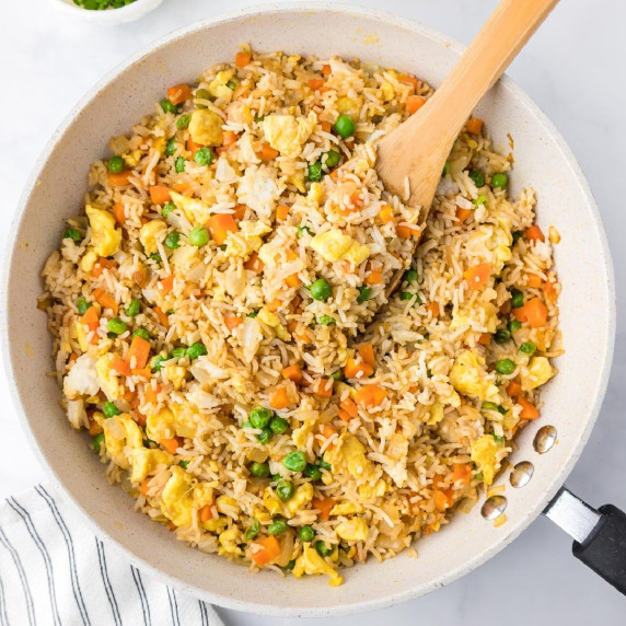 A skillet filled with fried rice mixed with peas, carrots, and scrambled eggs with a wooden spoon.