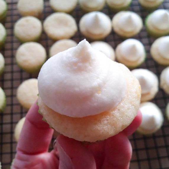 a mini cupcake topped with fluffy white frosting held above a cooling rack full of more cupcakes