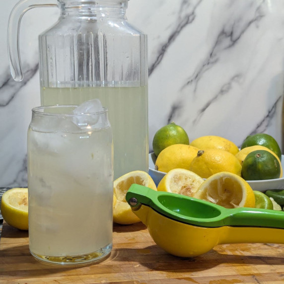 A glass and a pitcher of homemade lemonade with a juicer and lemons and limes in the background.
