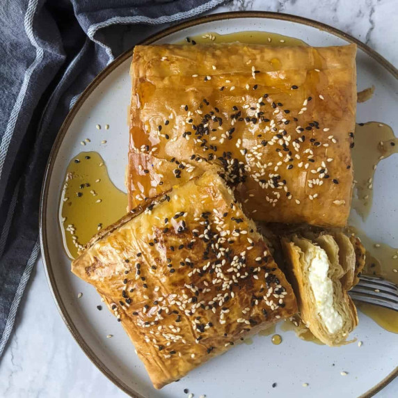 Two blocks of baked feta wrapped in filo served on plate with a bowl of honey and sesame seeds next 