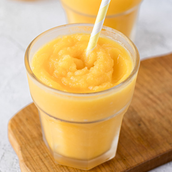 A glass of mango slushie with a straw on top of a wooden board.