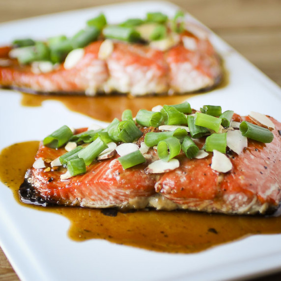 Side shot of two pieces of salmon garnished with green onions, almonds, and sauce on a white plate.