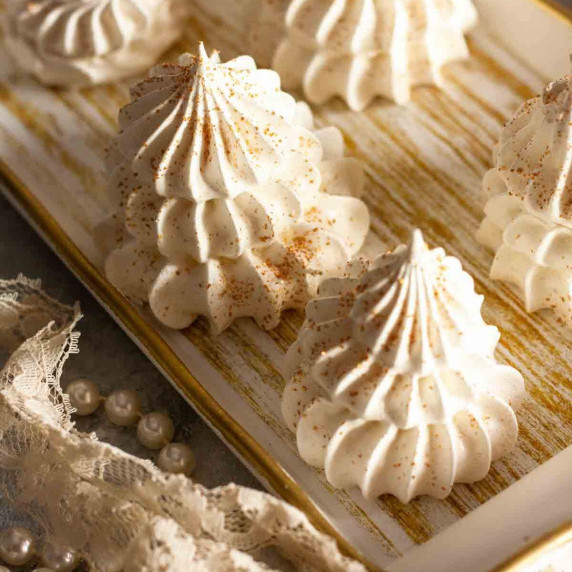 Meringue Christmas trees with edible gold glitter on a white and gold wooden tray next to lace.