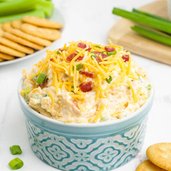 A blue and white ramekin piled high with million dollar dip topped with cheese, bacon, and scallions