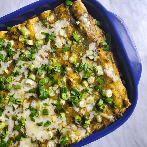 A shallow purple baking dish full of enchiladas dressed in melty cheese, green sauce, and onions.